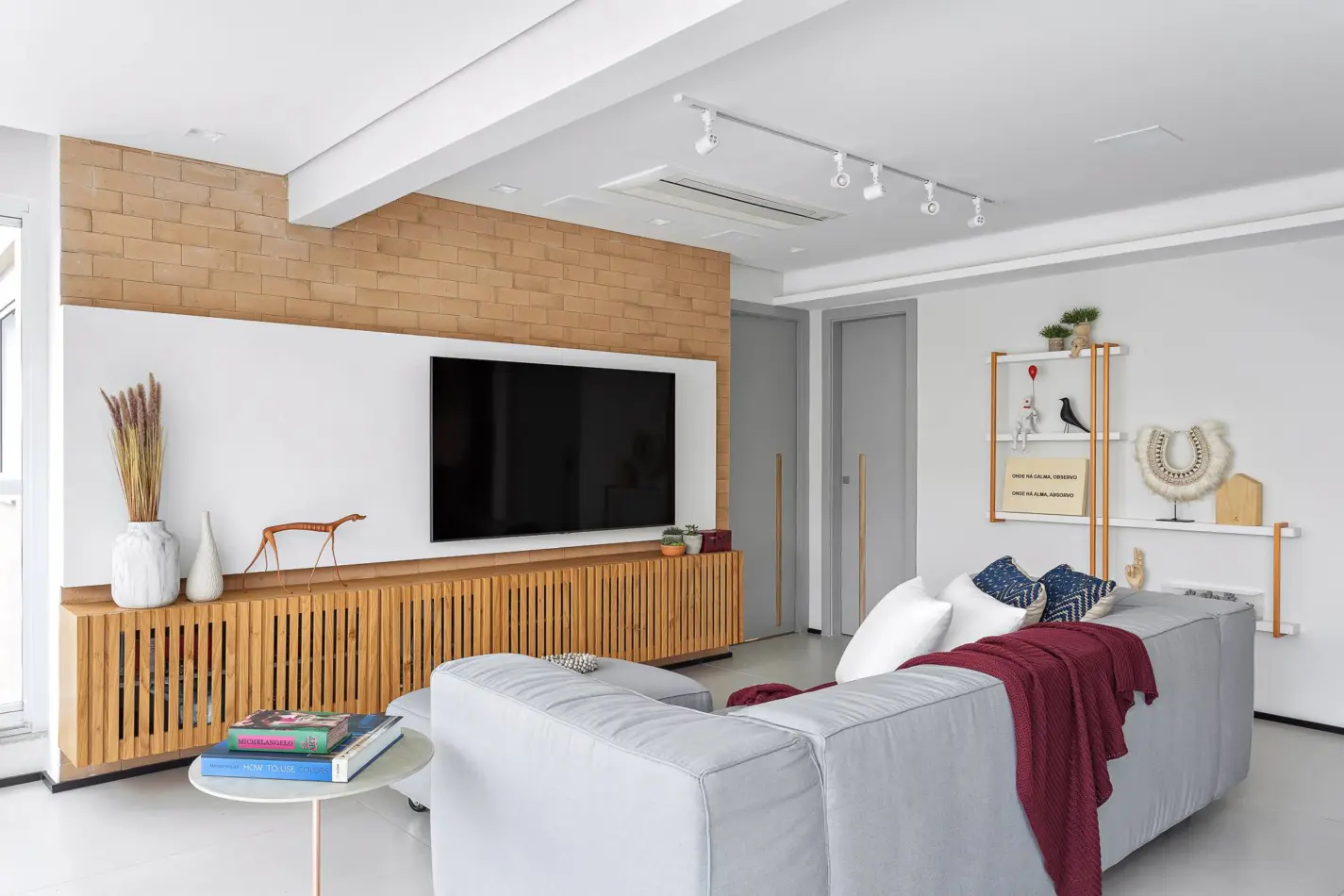 O que observar na hora de comprar o sofá para sua sala de estar. Projeto de Juliana Faria. Sala de estar moderna com sofá cinza, manta vinho, almofadas azuis e brancas. Rack de madeira ripada com TV preta, vasos decorativos e escultura de cachorro. Parede com tijolinhos e prateleiras brancas com objetos decorativos, como um colar de penas e um pássaro preto. Duas portas cinzas com puxadores de madeira ao fundo. Ambiente iluminado e aconchegante