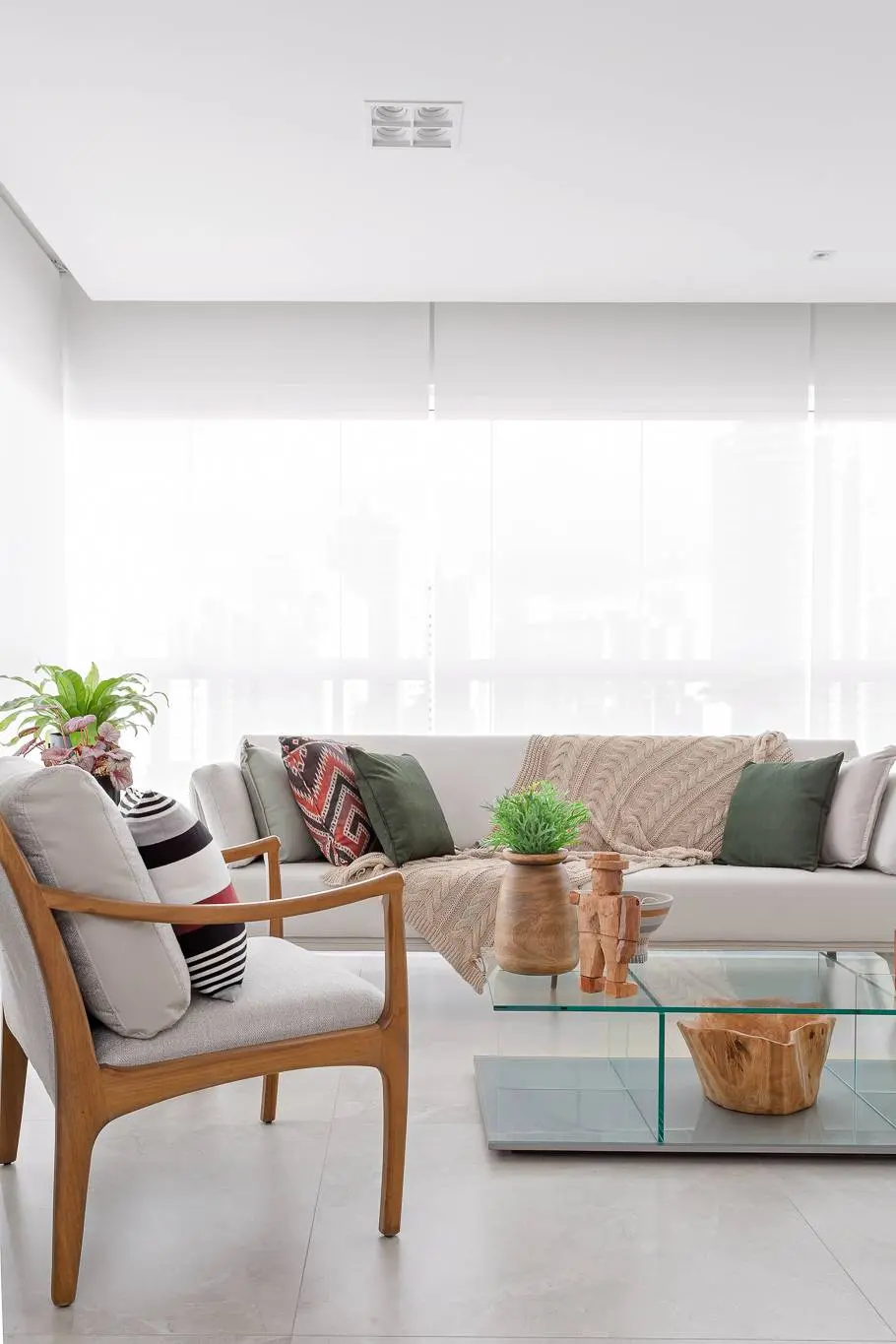 O que observar na hora de comprar o sofá para sua sala de estar. Projeto de Juliana Faria. Sala de estar clara com sofá branco, almofadas verdes e estampadas, manta bege. Uma poltrona de madeira clara e estofado cinza com almofada listrada. Mesa de centro de vidro com objetos decorativos de madeira e plantas. Cortinas brancas cobrem a janela ao fundo