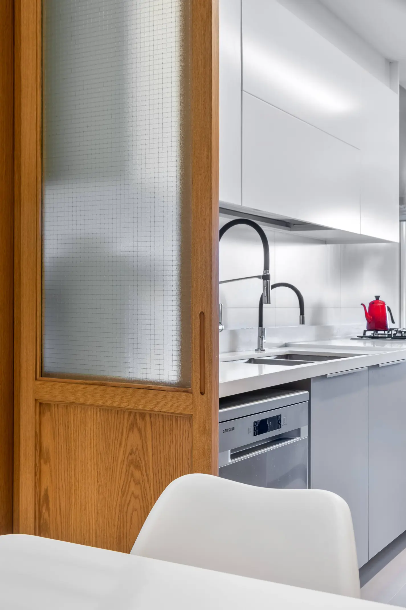 Apartamento iluminado e ventilado tem salinha de almoço e clima informal. Projeto de Sadala & Gomide Arquitetura. Cozinha moderna com porta de correr de madeira e vidro canelado à esquerda, revelando pia dupla com torneiras pretas, lava-louças Samsung e fogão com chaleira vermelha. Armários superiores brancos e inferiores cinzas. Uma cadeira branca aparece em primeiro plano à direita