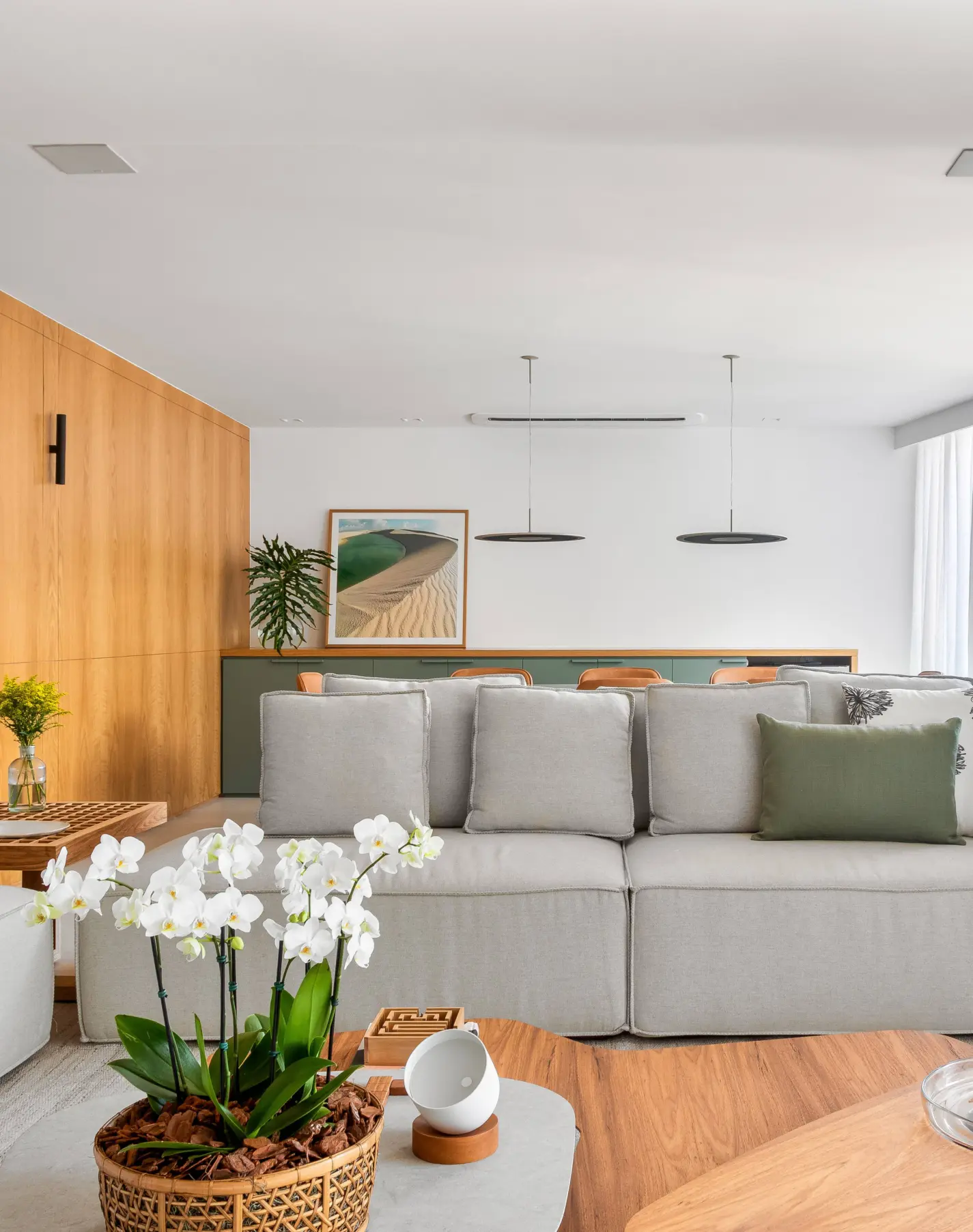 Apartamento iluminado e ventilado tem salinha de almoço e clima informal. Projeto de Sadala & Gomide Arquitetura. Sala de estar moderna com sofá modular cinza, almofada verde e orquídeas brancas em vaso de vime. Parede de madeira clara, armário verde e quadro de paisagem desértica.