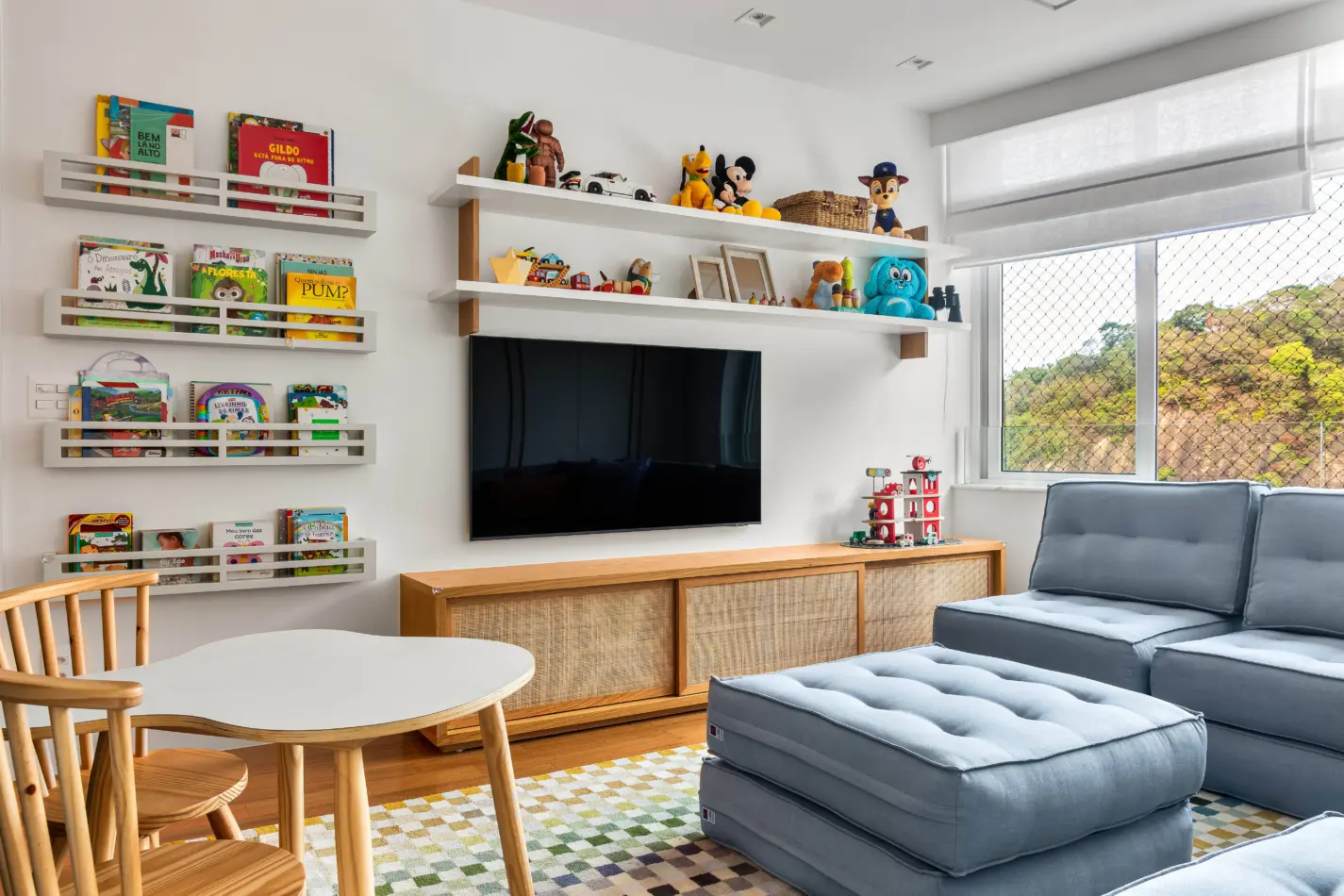 Apartamento iluminado e ventilado tem salinha de almoço e clima informal. Projeto de Sadala & Gomide Arquitetura. Sala de estar infantil com paredes brancas, estantes de livros e brinquedos, TV, mesa de madeira clara com cadeiras e sofá modular azul, vista para a natureza