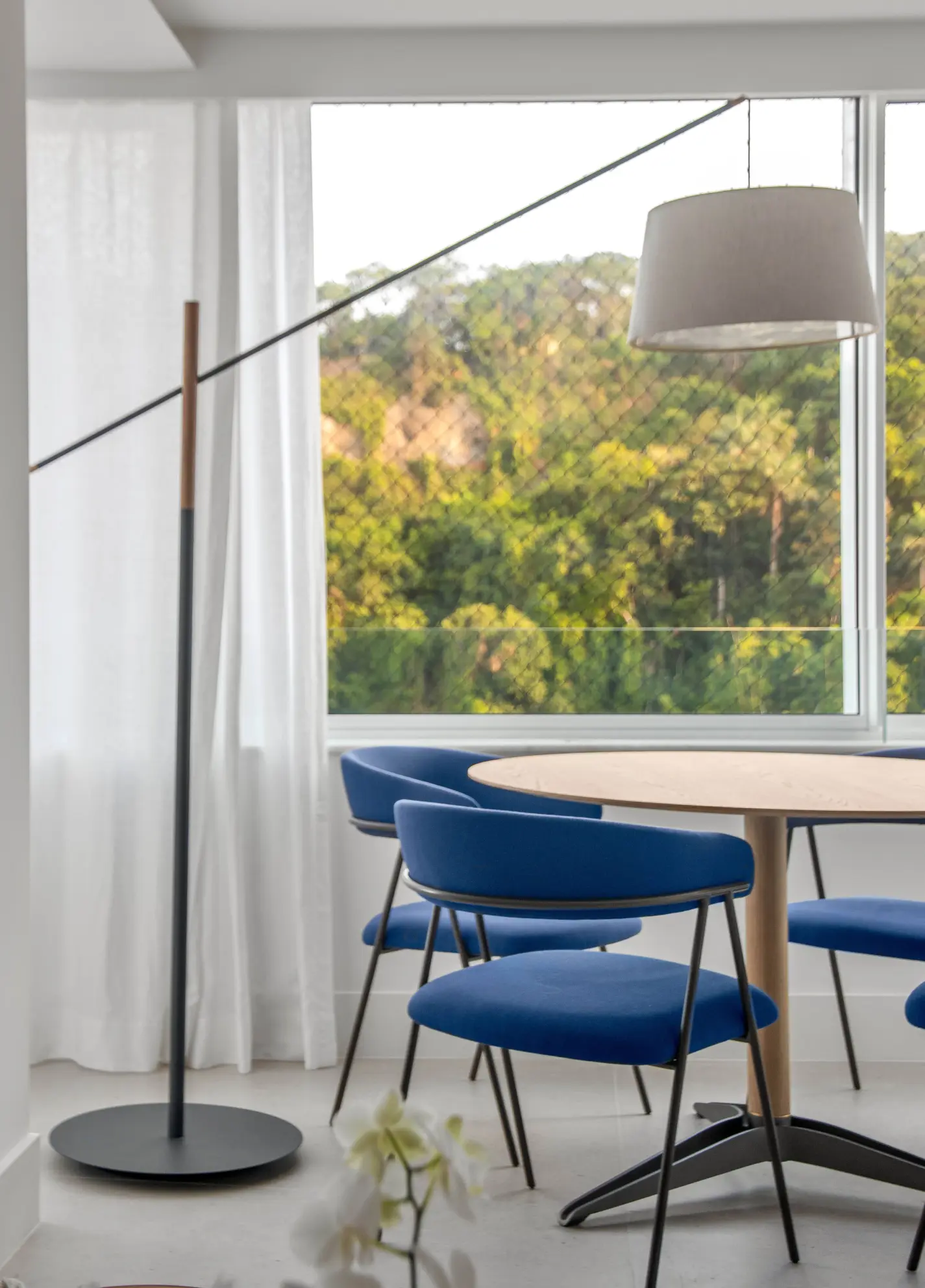 Apartamento iluminado e ventilado tem salinha de almoço e clima informal. Projeto de Sadala & Gomide Arquitetura. Sala de jantar com mesa redonda de madeira clara e quatro cadeiras azuis estofadas, sob uma luminária pendente cinza. À esquerda, um abajur de chão preto e madeira. Ao fundo, uma janela com cortina branca e vista para uma área verde com árvores