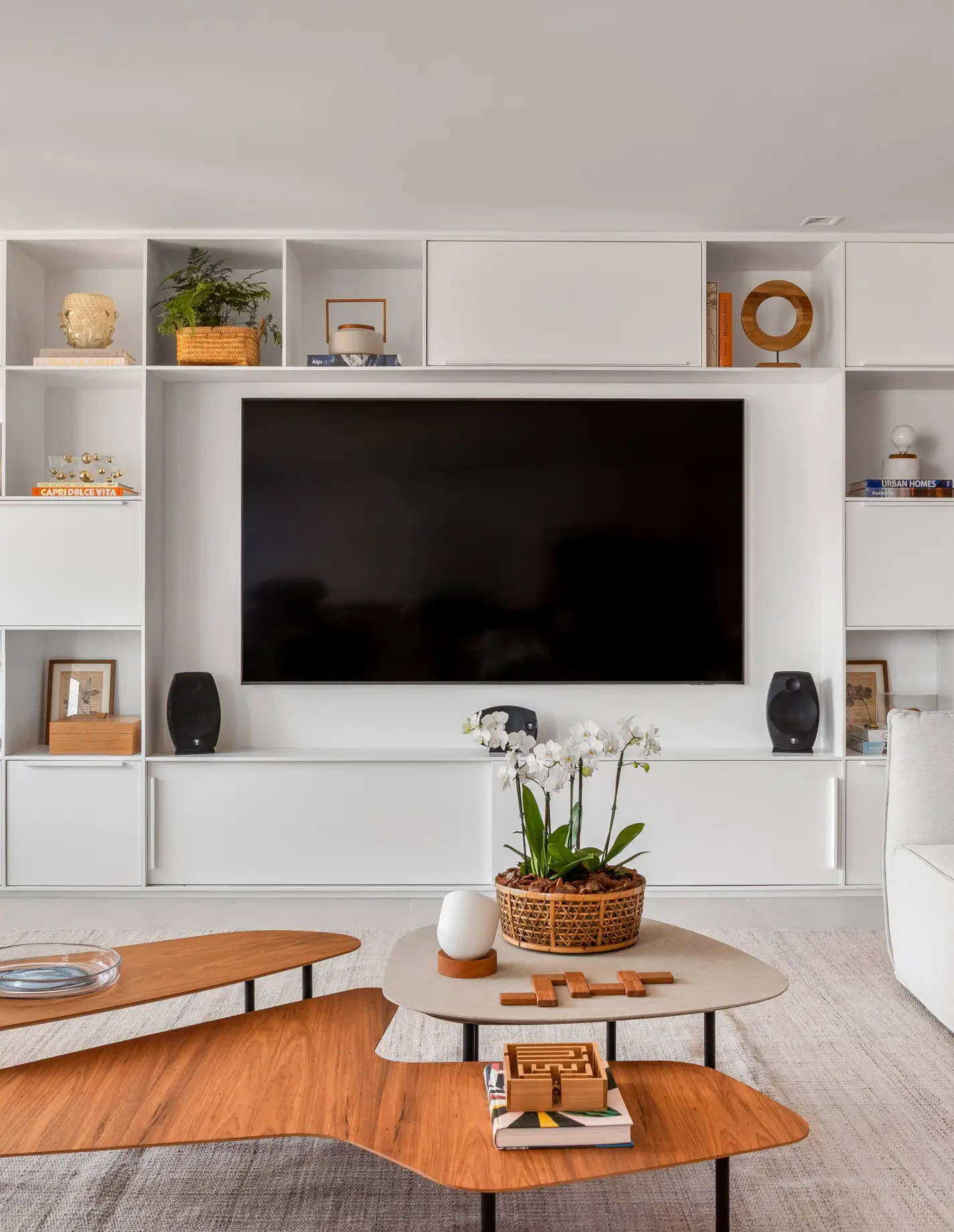 Apartamento iluminado e ventilado tem salinha de almoço e clima informal. Projeto de Sadala & Gomide Arquitetura. Sala de estar moderna com painel branco e televisão preta centralizada. Prateleiras abertas exibem livros, plantas e objetos decorativos. Duas mesas de centro de madeira com design orgânico, uma com orquídeas brancas em vaso de palha.