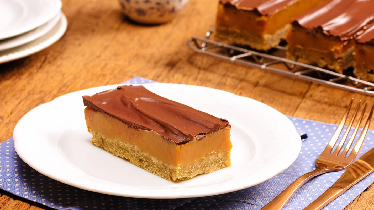 Fatia retangular de torta com base de biscoito, recheio de doce de leite e cobertura de chocolate, servida em prato branco sobre guardanapo azul de bolinhas brancas. Ao fundo, talheres dourados e outras fatias de torta em uma grade.